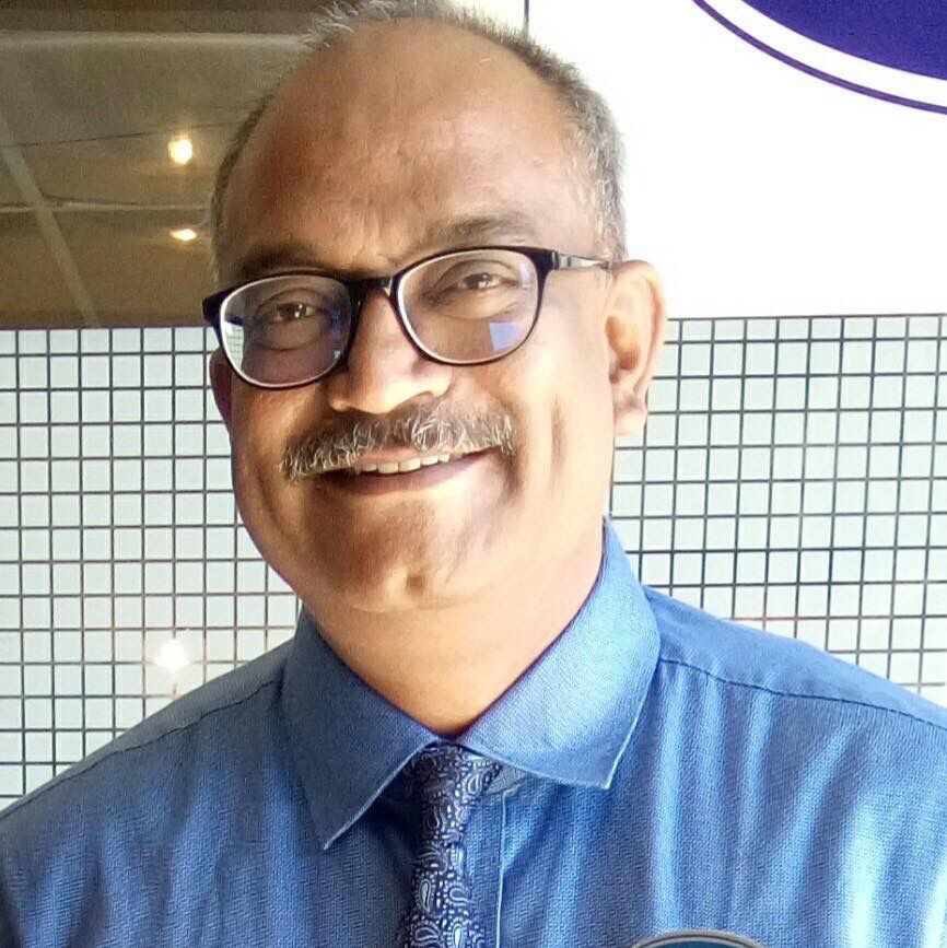 Professional close-up portrait of Nitin Aras smiling while wearing glasses, a blue button-down shirt, and a patterned tie
